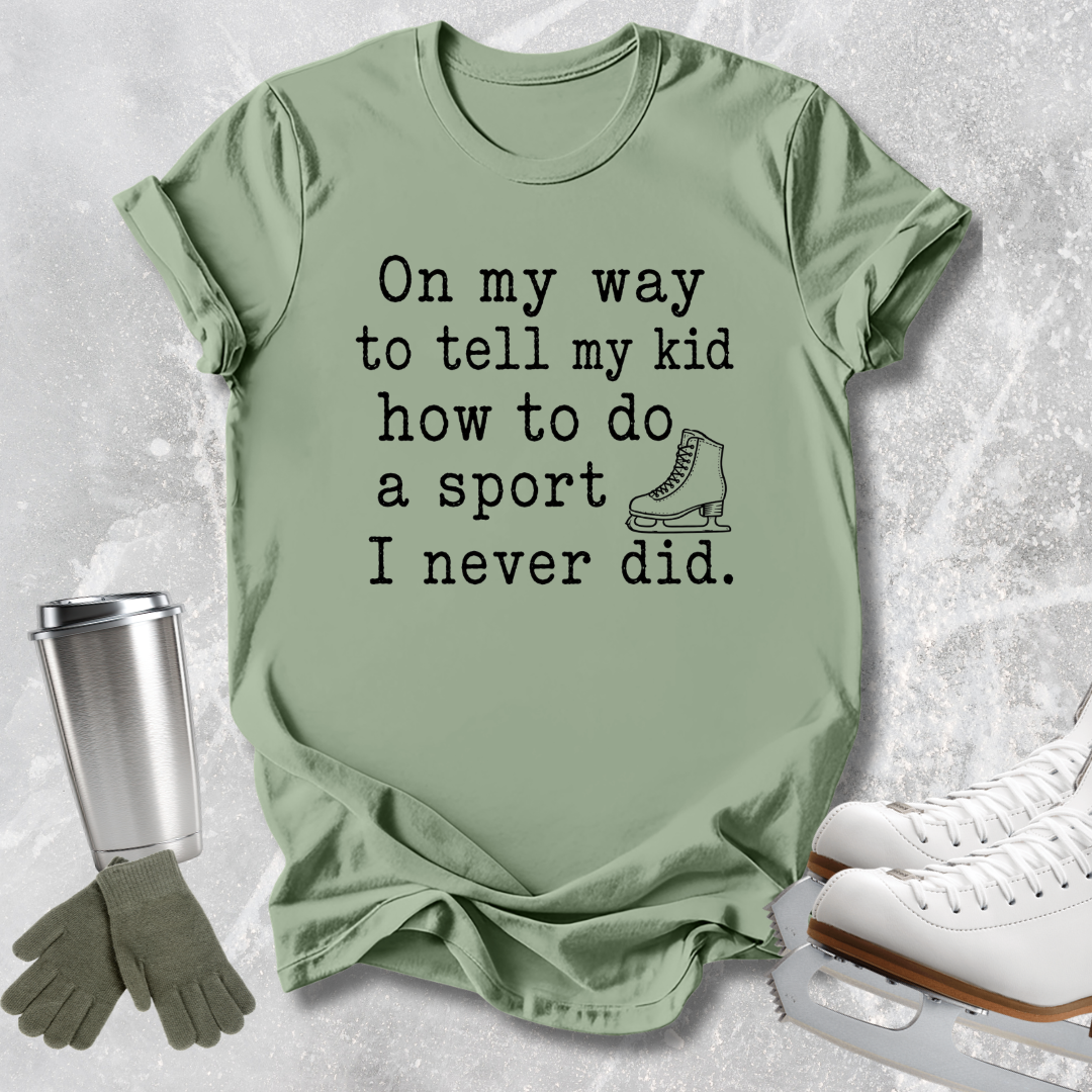 A sage green graphic t-shirt laid flat on a light gray textured background, styled with ice skates, a metal travel tumbler, and a pair of gloves. The t-shirt features black text that reads 'On my way to tell my kid how to do a sport I never did.