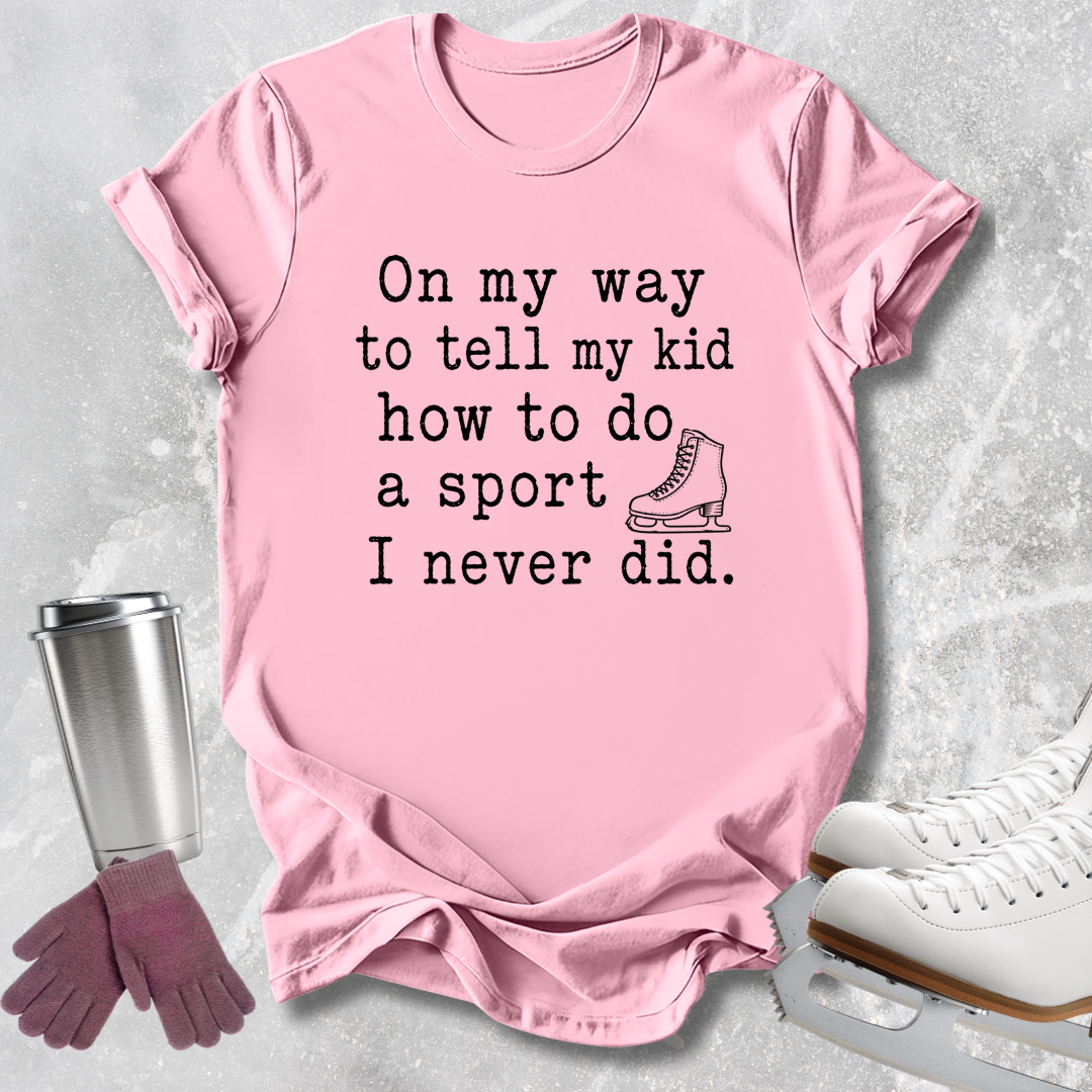 A light pink short-sleeve graphic T-shirt laid flat on a light gray textured surface, with black text reading 'On my way to tell my kid how to do a sport I never did.' and a small ice skate illustration.
