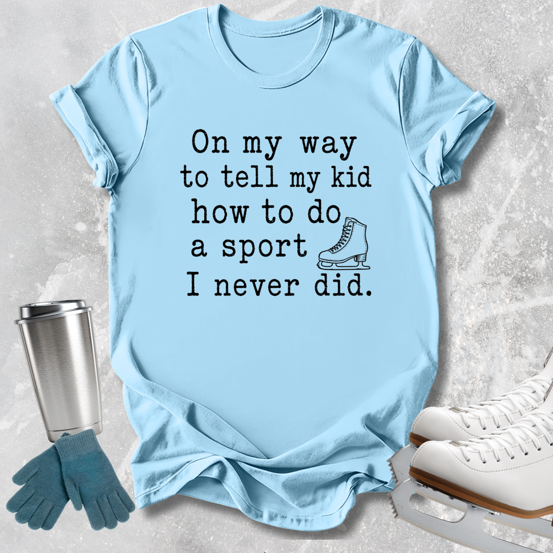 A light blue graphic T-shirt laid flat on a textured gray surface, with black text reading 'On my way to tell my kid how to do a sport I never did.' and a small ice skate illustration.