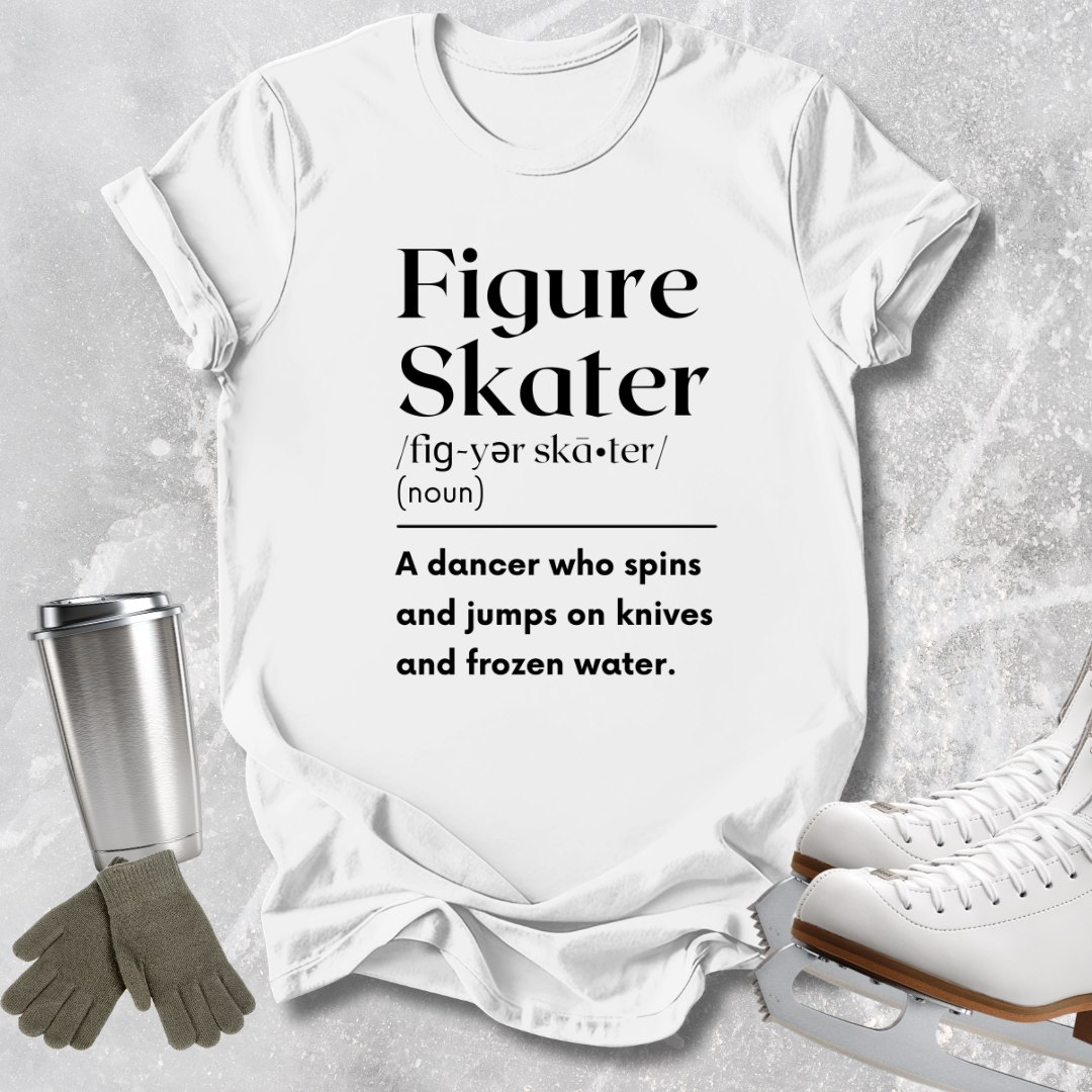 A white graphic T-shirt laid flat on a light gray textured surface, printed with the words 'Figure Skater' and a dictionary-style definition. Ice skates, gloves, and a metal cup are arranged around the shirt as styling props.