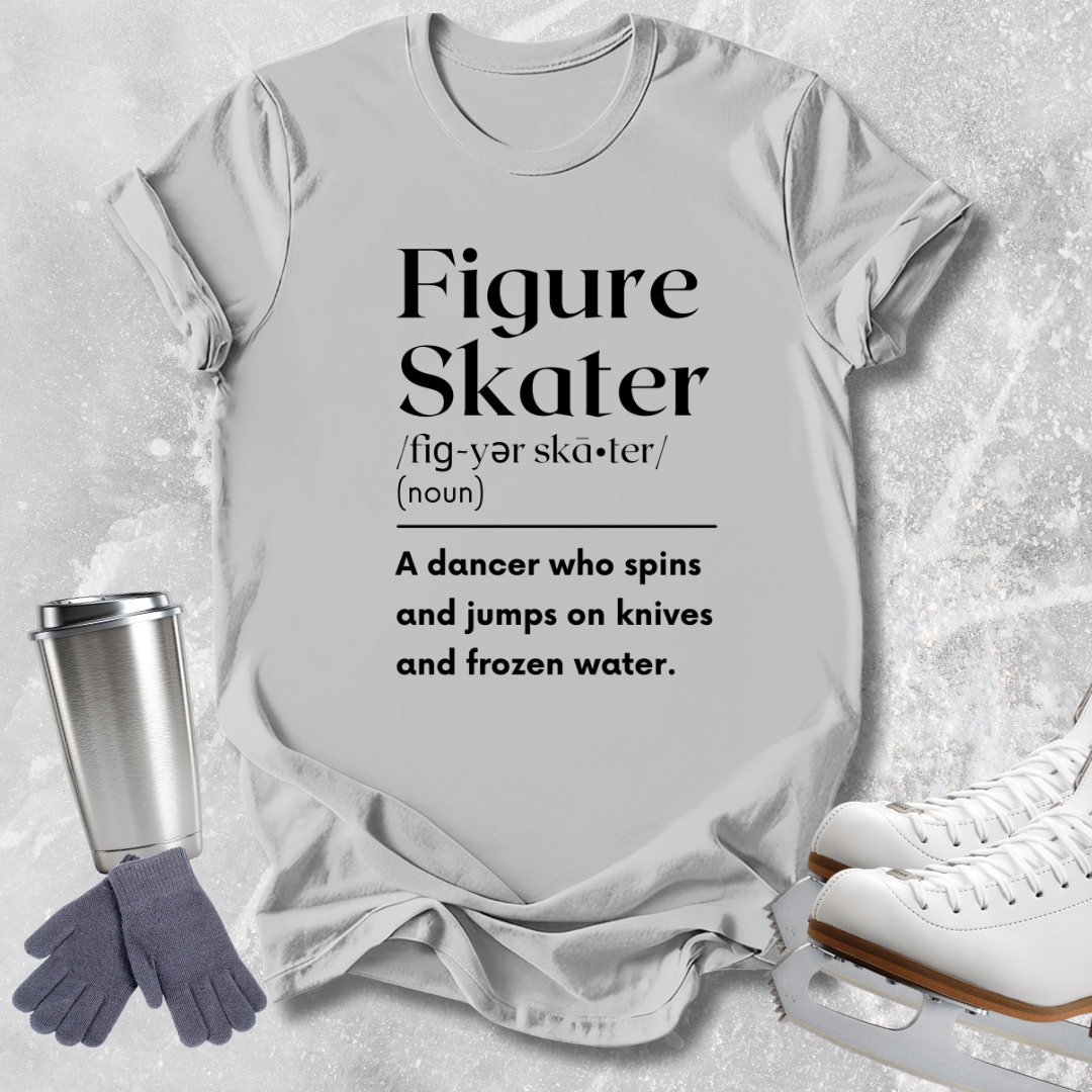 A light gray graphic T-shirt laid flat on a textured light gray surface, with black printed text defining the phrase 'Figure Skater.' Surrounding props include a stainless steel travel tumbler, gray gloves, and white ice skates.