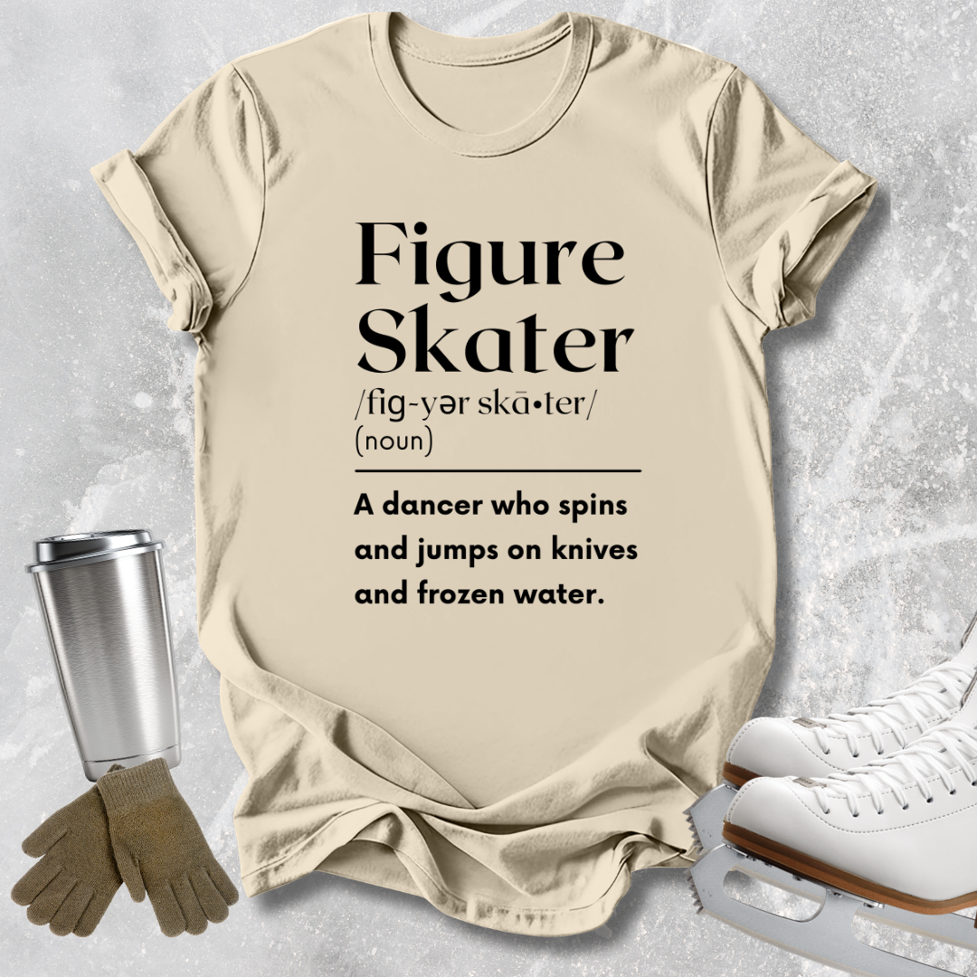 A beige graphic T-shirt laid flat on a light gray textured background, with printed black text reading 'Figure Skater' and a dictionary-style definition. Ice skates, gloves, and a metal tumbler are arranged around the shirt as styling props.
