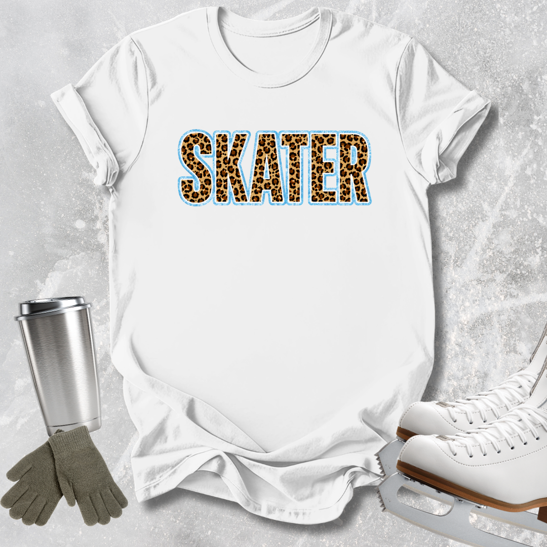 A white short-sleeve T-shirt laid flat on a light gray textured background, featuring the word "SKATER" in leopard-print letters with a blue outline. A stainless steel tumbler, gray gloves, and white ice skates are arranged around the shirt as styling props.