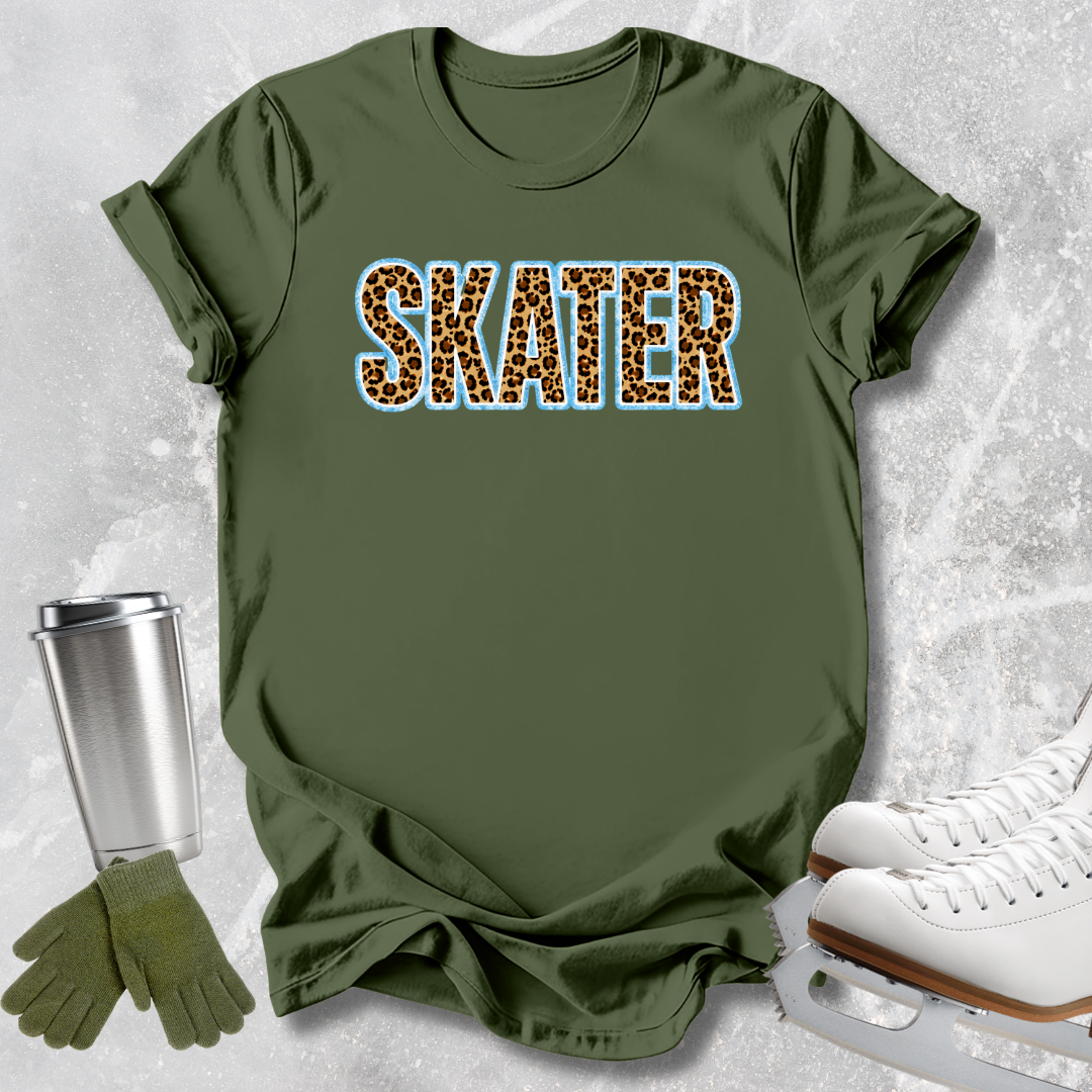 An olive green t-shirt with the word 'SKATER' printed across the chest in leopard print letters with a light blue outline. The shirt is styled in a flat-lay arrangement with ice skates, a metal travel mug, and a pair of green gloves on a textured gray background.