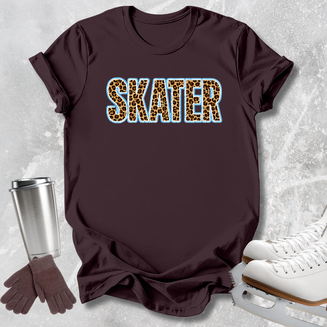 A dark brown short-sleeve t-shirt with the word 'SKATER' printed across the chest in leopard-print letters outlined in light blue. The shirt is styled in a flat lay with white ice skates, a silver travel mug, and brown gloves on a light gray textured background.
