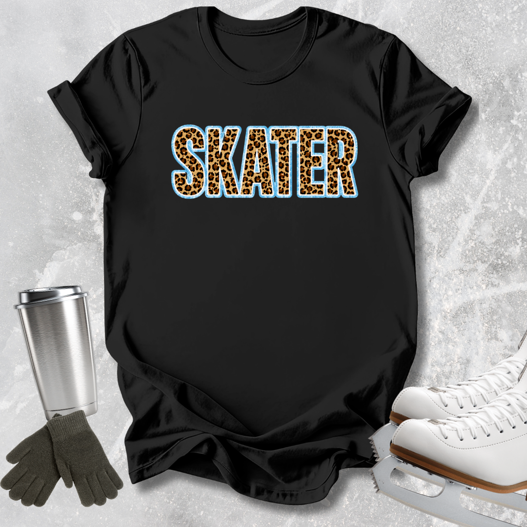 A black short-sleeve T-shirt laid flat on a light textured background, featuring the word "SKATER" in leopard-print letters with a blue outline. White ice skates, dark gloves, and a metal cup are arranged around the shirt as styling props.