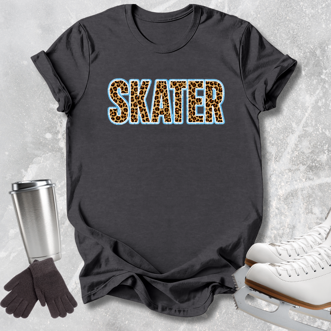 A dark charcoal gray t-shirt with the word 'SKATER' printed across the chest in a leopard print pattern with a light blue outline. The shirt is styled in a flat-lay arrangement on a textured gray background, accompanied by white ice skates, a silver travel mug, and a pair of dark gray gloves.