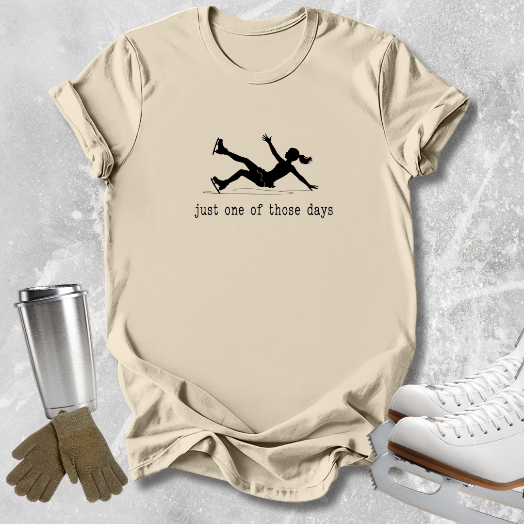 A beige graphic t-shirt laid flat on a light gray textured surface, featuring a black silhouette of a female ice skater falling with the text 'just one of those days' underneath. The shirt is styled with ice skates, a metal travel tumbler, and a pair of gloves.