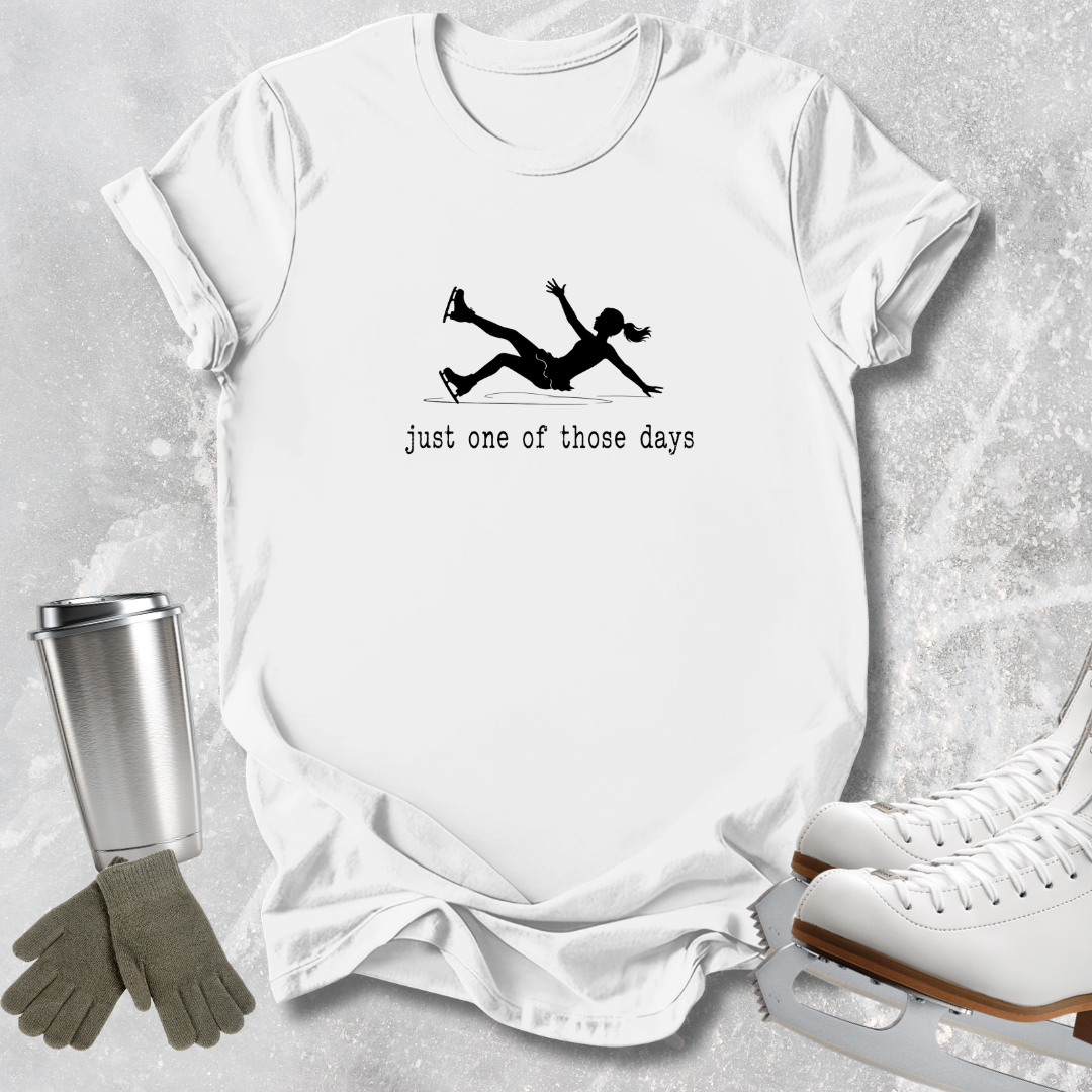 A white short-sleeve graphic T-shirt laid flat on a light gray textured surface, with a black silhouette of a person ice skating and the text 'just one of those days' printed on the front. A stainless steel cup, gray gloves, and white ice skates are arranged around the shirt as styling props.