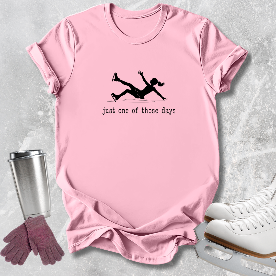 A light pink short-sleeve t-shirt with a black silhouette of a person falling and the text 'just one of those days' printed on the front. The shirt is displayed in a flat-lay arrangement on a textured gray background, accompanied by ice skates, gloves, and a travel tumbler.