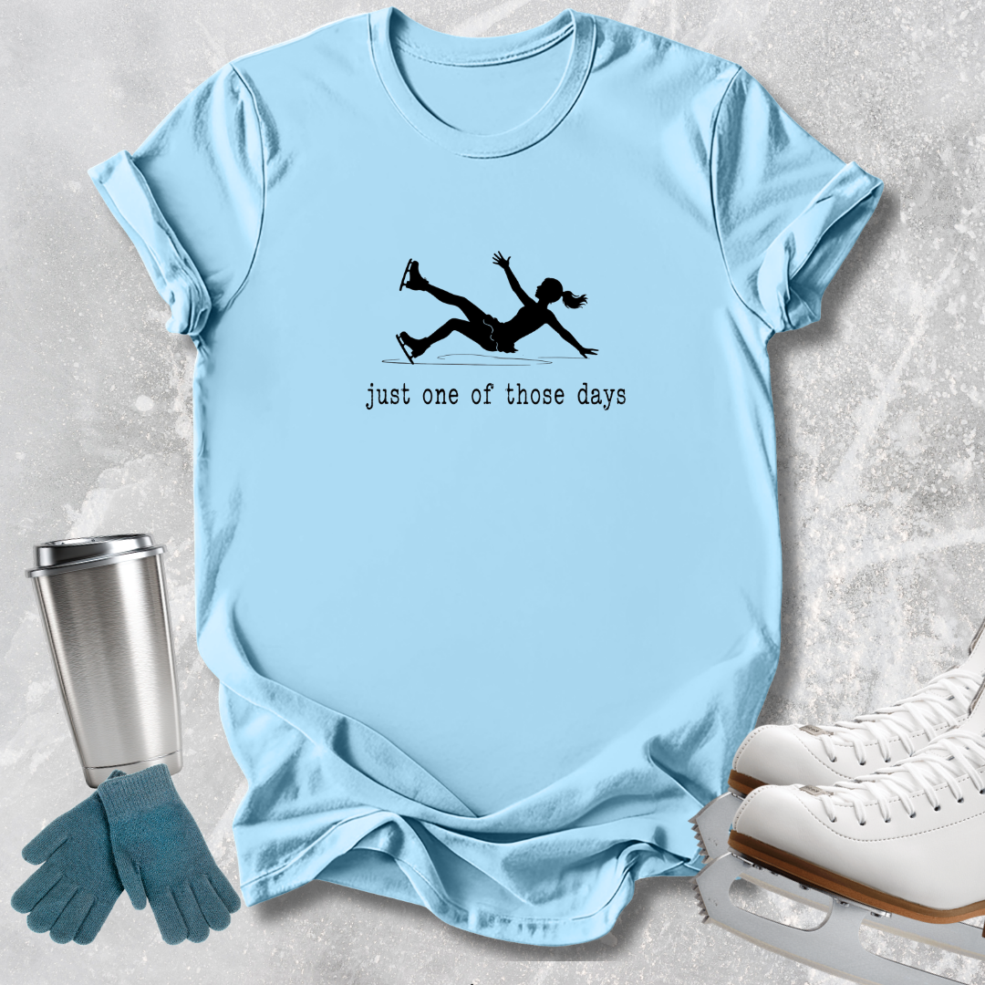 A light blue graphic t-shirt laid flat on a textured gray background, featuring a black silhouette of an ice skater and the text 'just one of those days'. The shirt is styled with ice skates, a metal travel mug, and blue gloves.
