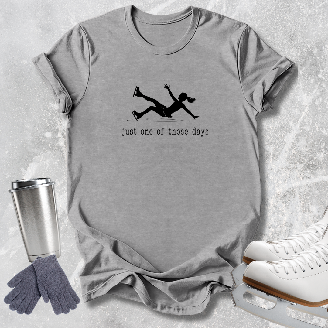 A heather gray short-sleeve t-shirt with a black graphic of an ice skater and the text 'just one of those days', styled in a flat lay with white ice skates, a silver travel mug, and gray gloves on a light gray textured background.