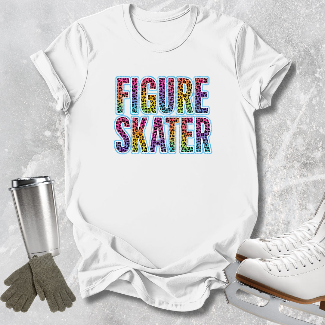 A white short-sleeve T-shirt laid flat on a light gray textured surface, featuring the words 'FIGURE SKATER' in large multicolor letters. A stainless steel tumbler, gray gloves, and white ice skates are arranged around the shirt as styled props.