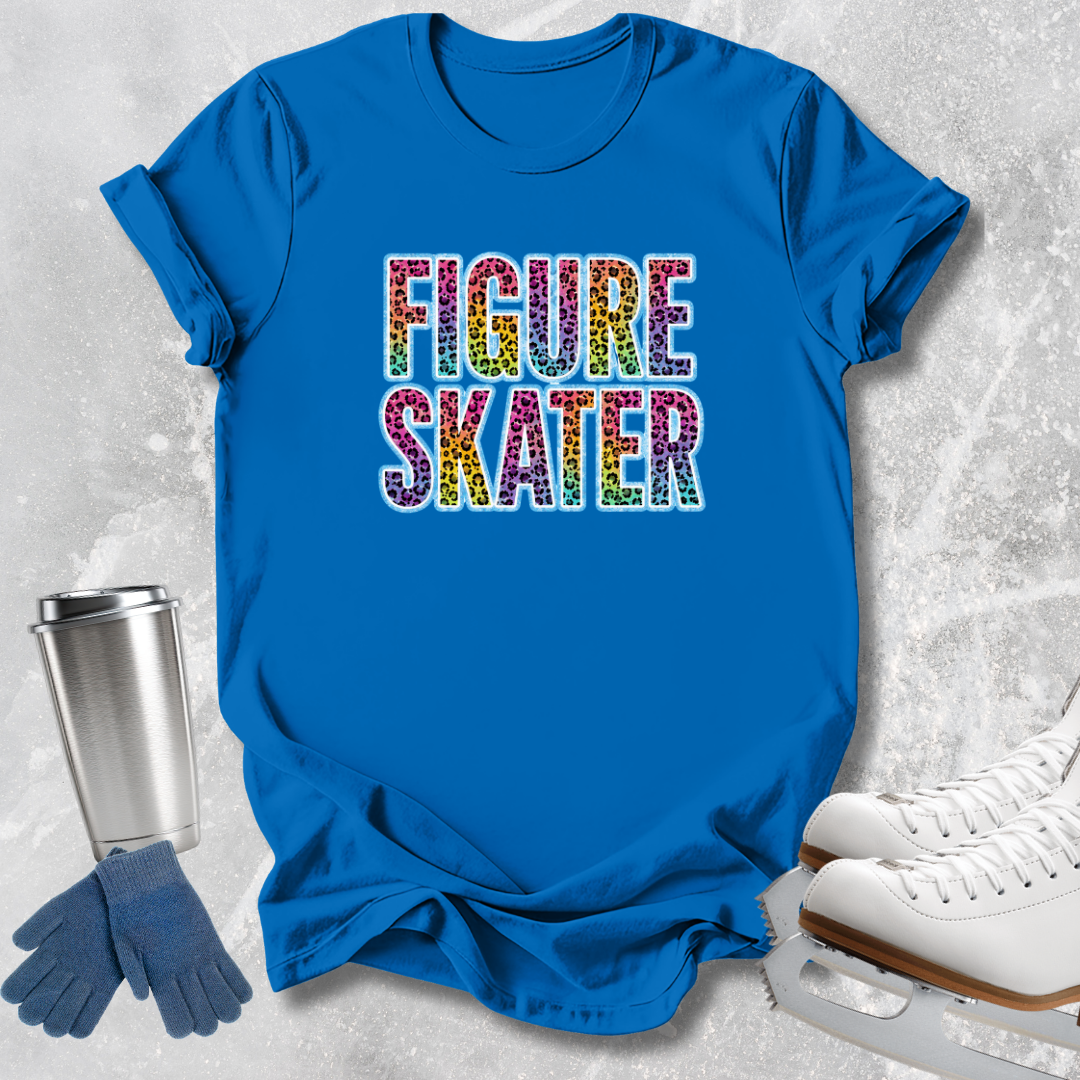 A bright blue graphic T-shirt laid flat on a light textured surface, with the words "FIGURE SKATER" printed across the chest in multicolored glitter-style lettering. Blue gloves, a silver tumbler, and white ice skates are arranged around the shirt as styling props.