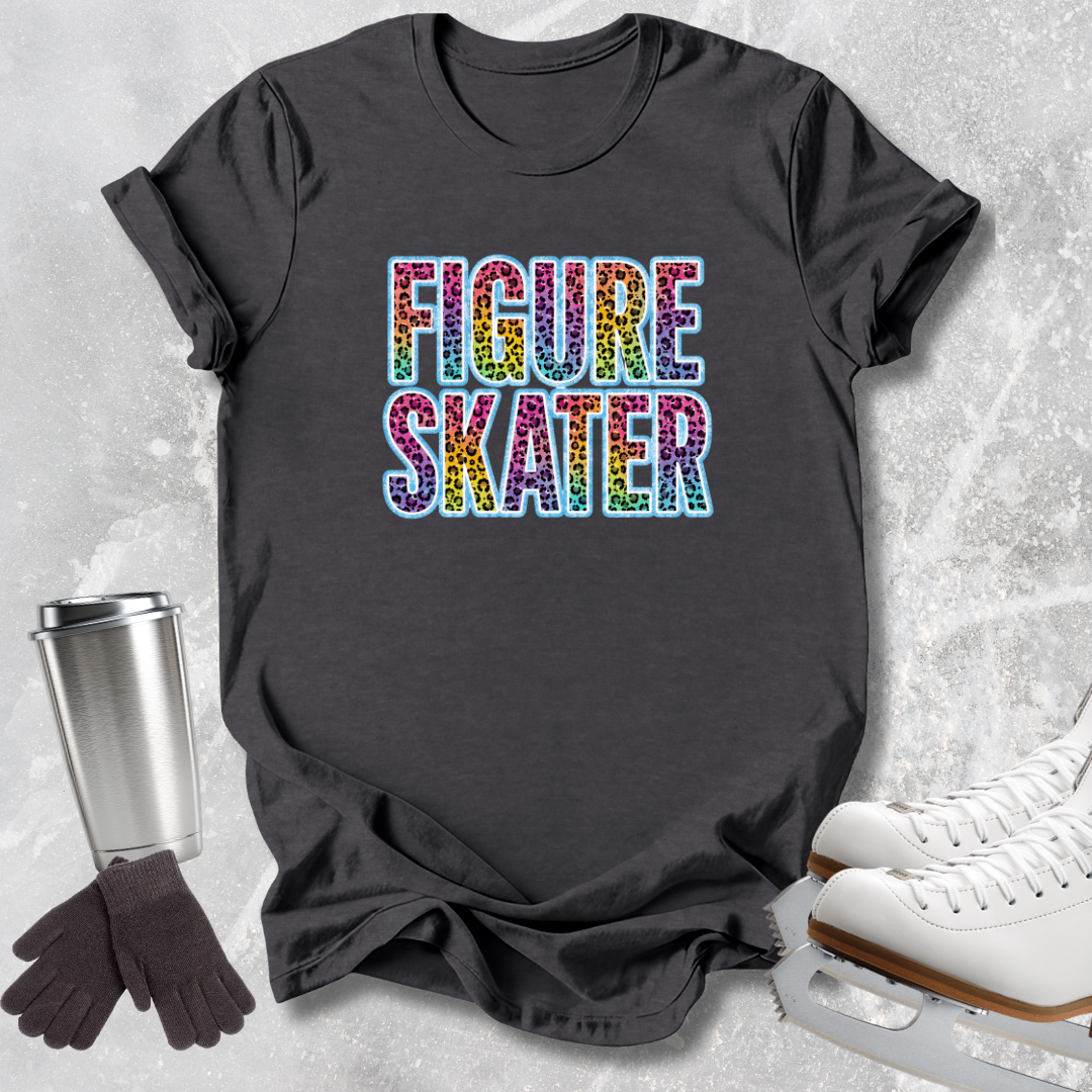 A dark charcoal gray graphic t-shirt featuring the words 'FIGURE SKATER' in a colorful, glittery rainbow font. The shirt is styled in a flat-lay arrangement on a textured light gray background, accompanied by white ice skates, a stainless steel travel mug, and a pair of dark gray gloves.