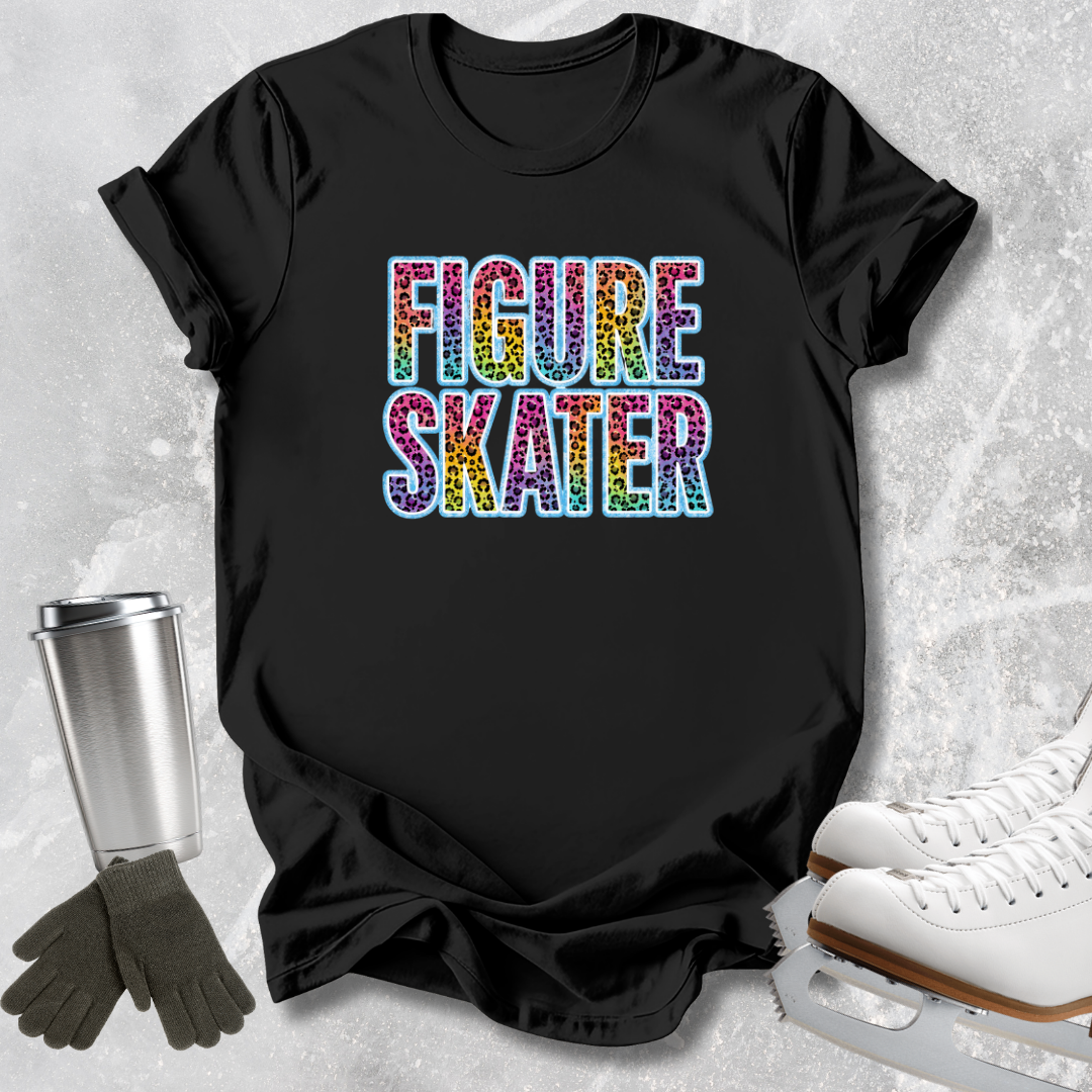 A black graphic t-shirt featuring the words 'FIGURE SKATER' in a colorful, glittery rainbow font. The shirt is presented in a flat-lay arrangement alongside ice skate boots, gloves, and a metal travel mug on a textured light gray background.