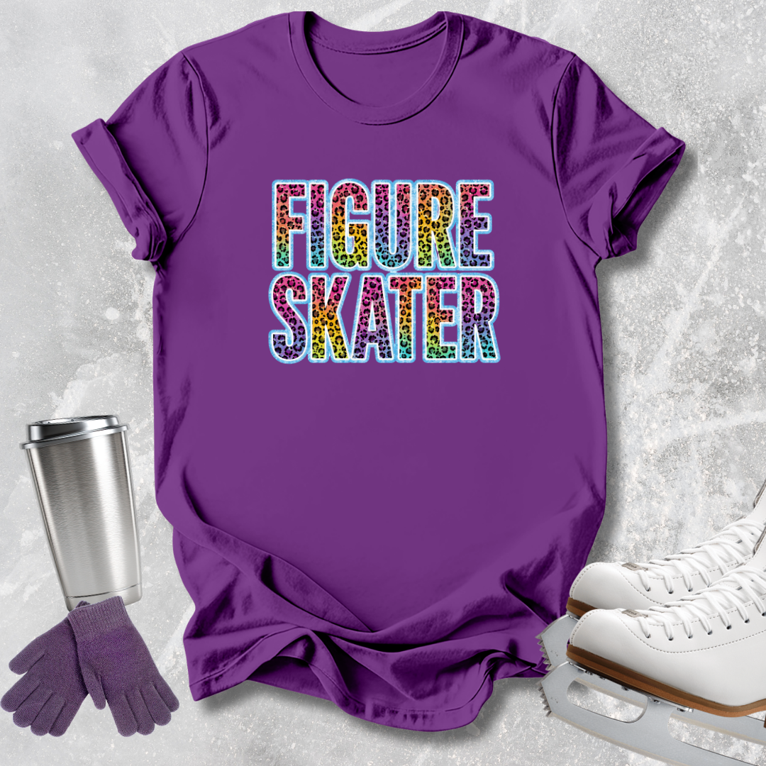 A purple short-sleeve graphic T-shirt laid flat on a light gray textured surface, with the words "FIGURE SKATER" printed across the chest in rainbow-colored letters. White ice skates, purple gloves, and a stainless steel travel mug are arranged around the shirt as styling props.