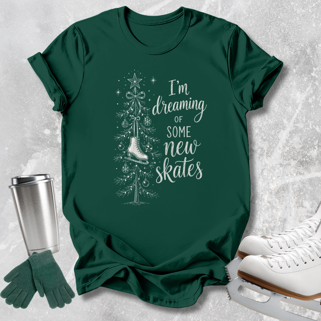 A dark green graphic t-shirt featuring a white illustration of a Christmas tree and ice skate with the text 'I'm dreaming of some new skates'. The shirt is styled in a flat-lay arrangement on a textured gray background, accompanied by a silver travel mug, green knit gloves, and white ice skates.