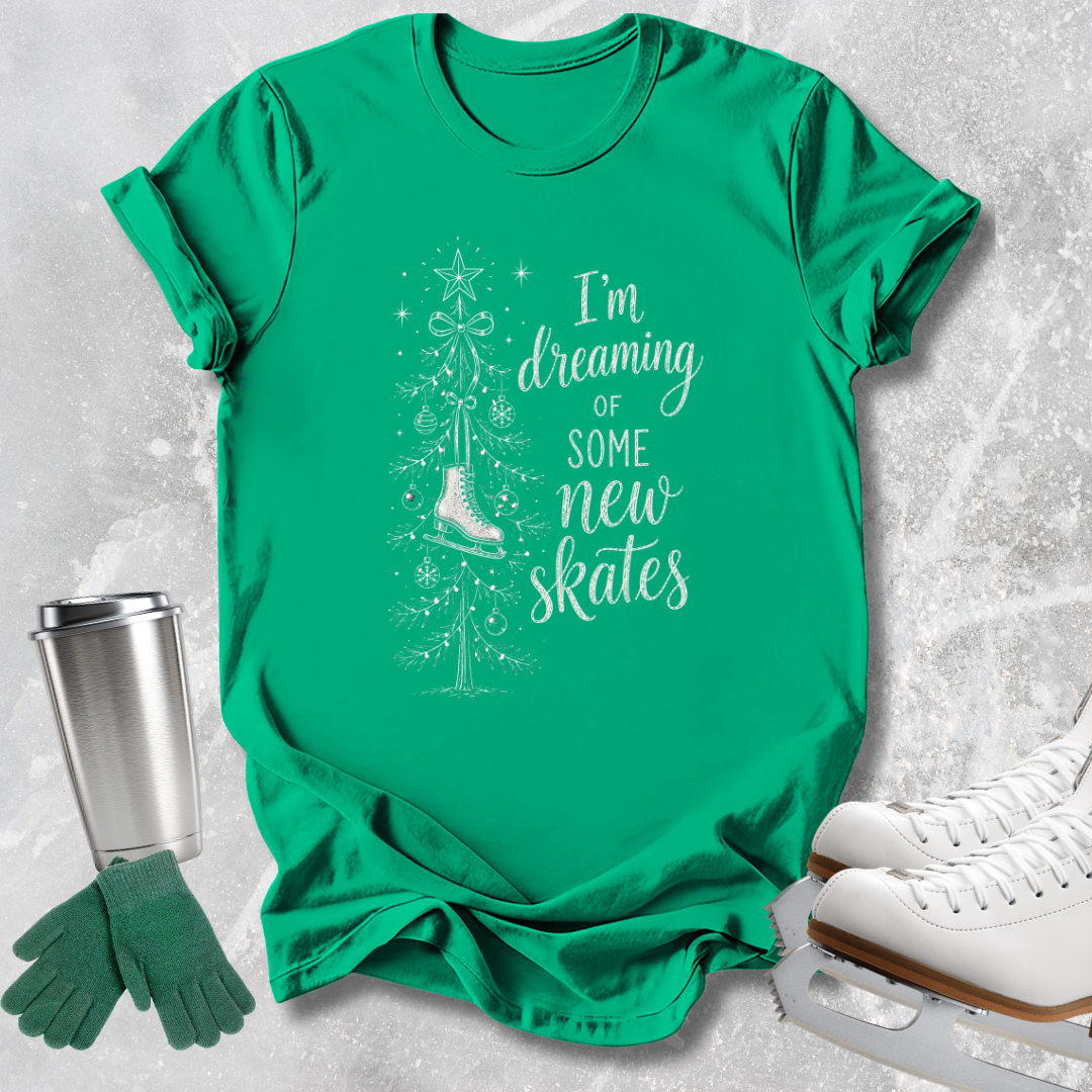 A bright green short-sleeve T-shirt laid flat on a light gray textured surface, featuring a white graphic of a Christmas tree, hanging ornament, ice skate, and the phrase 'I'm dreaming of some new skates.' Green knit gloves and white ice skates appear as styled props around the shirt.