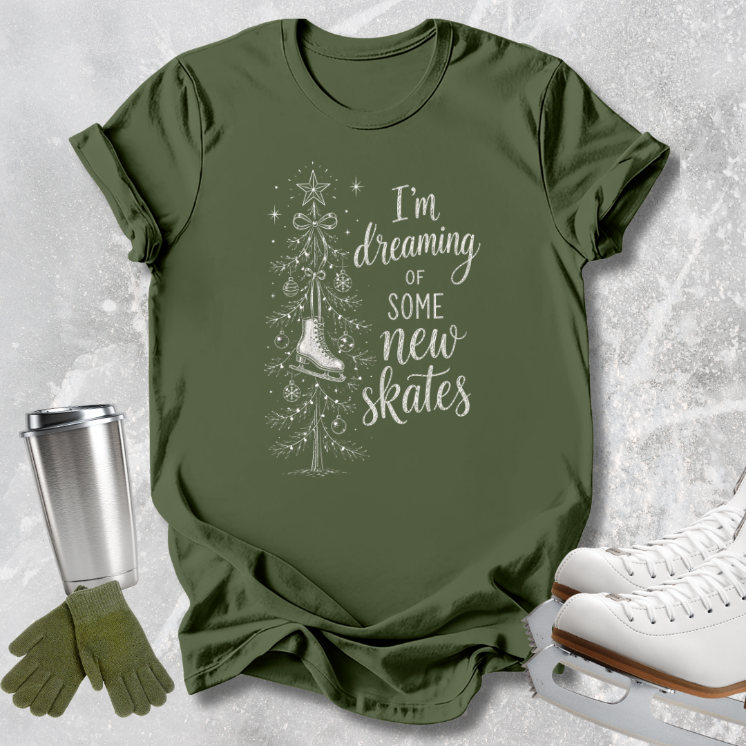 An olive green graphic t-shirt laid flat on a light gray textured background, styled with a stainless steel travel mug, green knit gloves, and white ice skates. The t-shirt features a white graphic of a Christmas tree and a single ice skate with the text 'I'm dreaming of some new skates'.