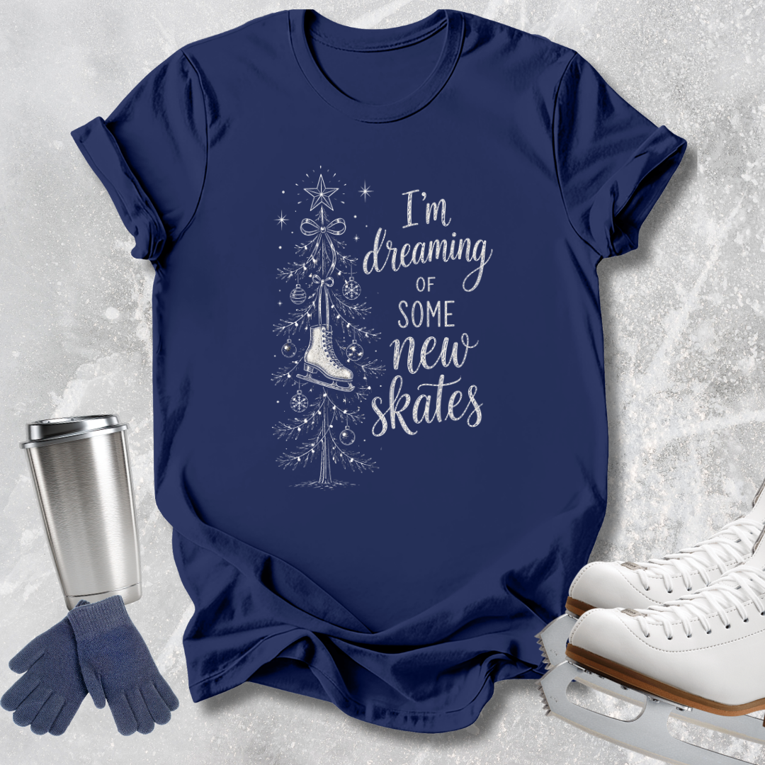 A navy blue short-sleeve graphic T-shirt laid flat on a light textured surface, with a white illustration of a Christmas tree, a skating boot, and the phrase 'I'm dreaming of some new skates.' White ice skates, blue gloves, and a metal tumbler are arranged around the shirt as styling props.