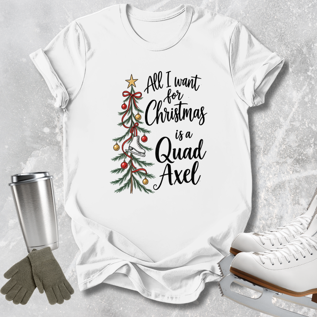 A white graphic t-shirt laid flat on a light gray textured surface, featuring a Christmas tree illustration and the phrase 'All I want for Christmas is a Quad Axel' in black script. A stainless steel tumbler, gray gloves, and white ice skates are arranged around the shirt as styled props.
