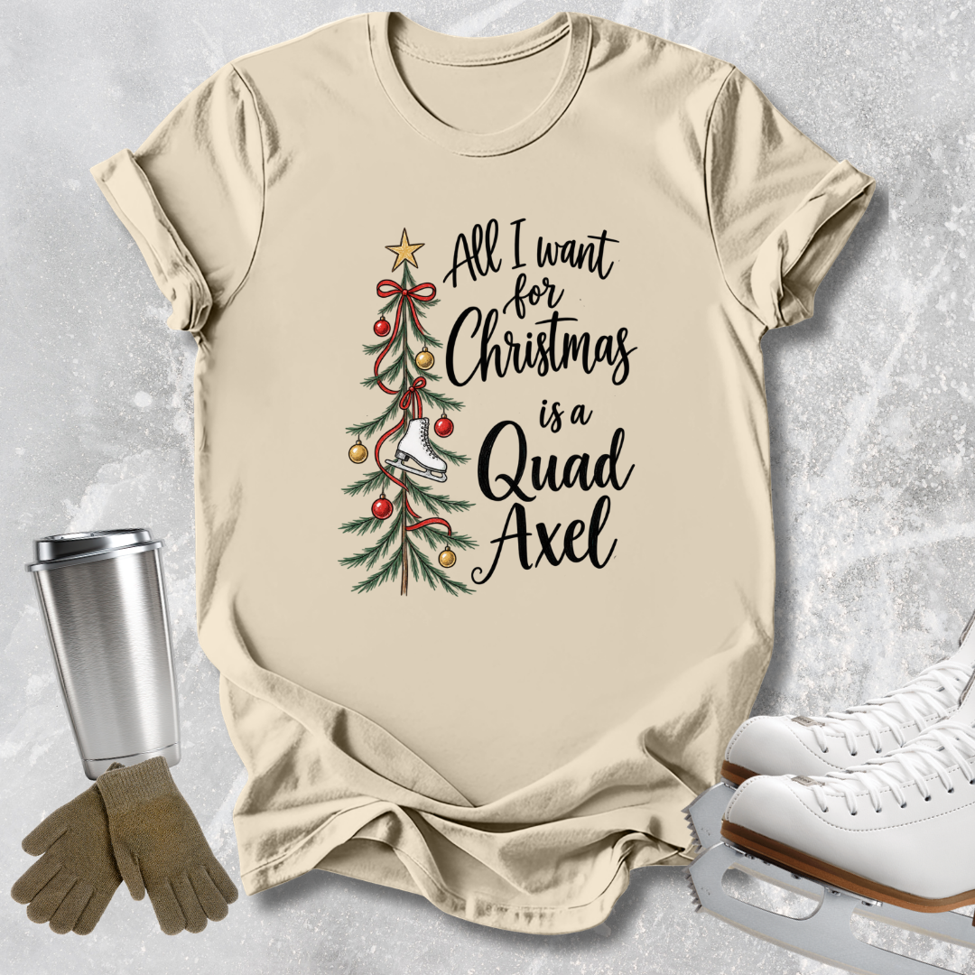 A beige graphic T-shirt laid flat on a light gray textured surface, with a Christmas tree illustration and the phrase 'All I want for Christmas is a Quad Axel' printed on the front. White ice skates, tan gloves, and a metal tumbler are placed around it as styling props.