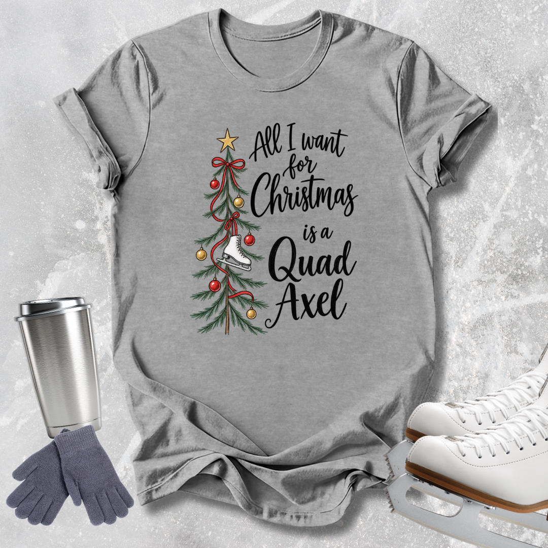 A heather gray graphic t-shirt featuring a Christmas tree illustration and the text 'All I want for Christmas is a Quad Axel' in black script. The shirt is styled in a flat-lay arrangement on a textured light gray background, accompanied by a silver travel mug, gray gloves, and white ice skates.