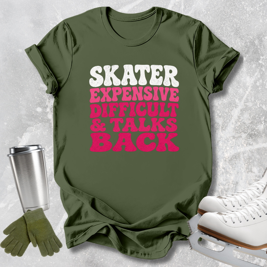 An olive green graphic t-shirt laid flat on a light gray textured background, styled with ice skates, a metal travel tumbler, and a pair of gloves. The t-shirt features the text 'SKATER EXPENSIVE DIFFICULT & TALKS BACK' in white and bright pink lettering.