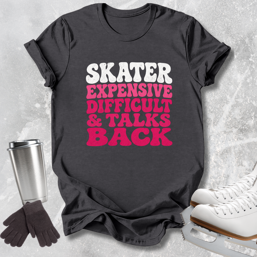 A dark charcoal gray graphic t-shirt laid flat on a light gray textured surface, surrounded by skating-themed props including white ice skates, a stainless steel travel mug, and a pair of black gloves. The t-shirt features the text 'SKATER EXPENSIVE DIFFICULT & TALKS BACK' in white and bright pink lettering.