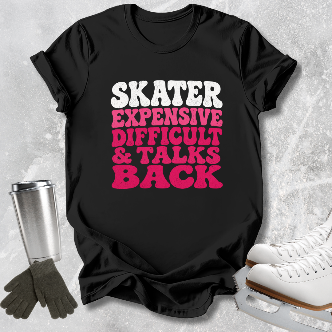 A black graphic t-shirt with the text 'SKATER EXPENSIVE DIFFICULT & TALKS BACK' in white and pink bubble letters, styled in a flat-lay with ice skates, gloves, and a travel tumbler on a light gray textured background.