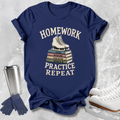 A navy blue graphic T-shirt laid flat on a light gray textured surface, featuring the words 'HOMEWORK PRACTICE REPEAT' in cream around an illustration of white ice skates stacked on books. A stainless steel tumbler, blue gloves, and white ice skates are arranged around the shirt as styled props.