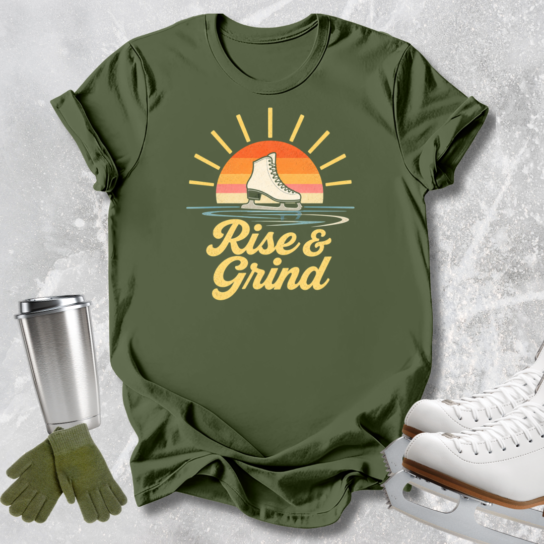 An olive green graphic t-shirt laid flat on a light gray textured surface, styled with ice skates, a metal travel tumbler, and a pair of gloves. The t-shirt features a retro sunset graphic with a single ice skate and the text 'Rise & Grind'.