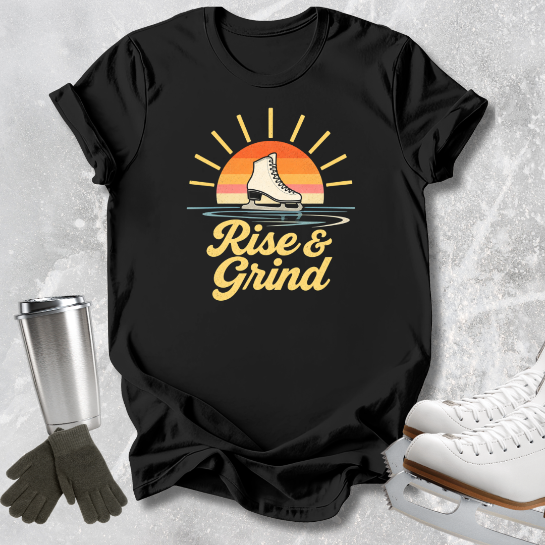 A black graphic t-shirt laid flat on a light gray textured surface, featuring a retro sunset design with a hockey skate and the words 'Rise & Grind' in yellow. White ice skates, dark gloves, and a metal tumbler are styled around the shirt.