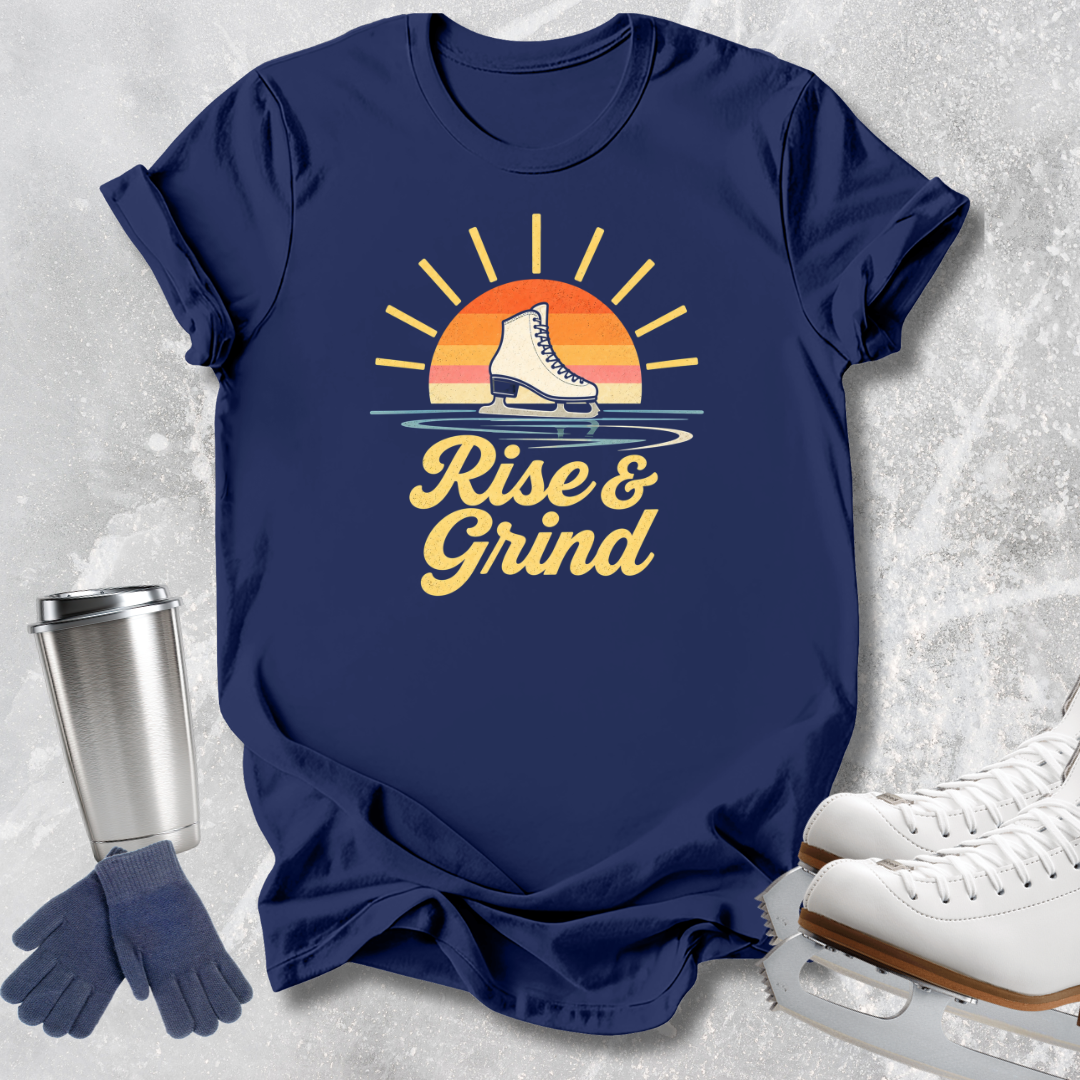 A navy blue graphic T-shirt laid flat on a light textured surface, featuring a retro sunset and ice skate design with the words 'Rise & Grind.' White ice skates, blue gloves, and a metal travel tumbler are arranged around the shirt as styling props.