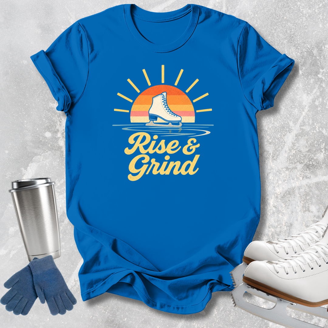 A bright blue graphic T-shirt laid flat on a light textured surface, featuring a retro sunset and ice skate design with the words 'Rise & Grind.' White ice skates, blue gloves, and a stainless steel travel mug are arranged around the shirt as styled props.