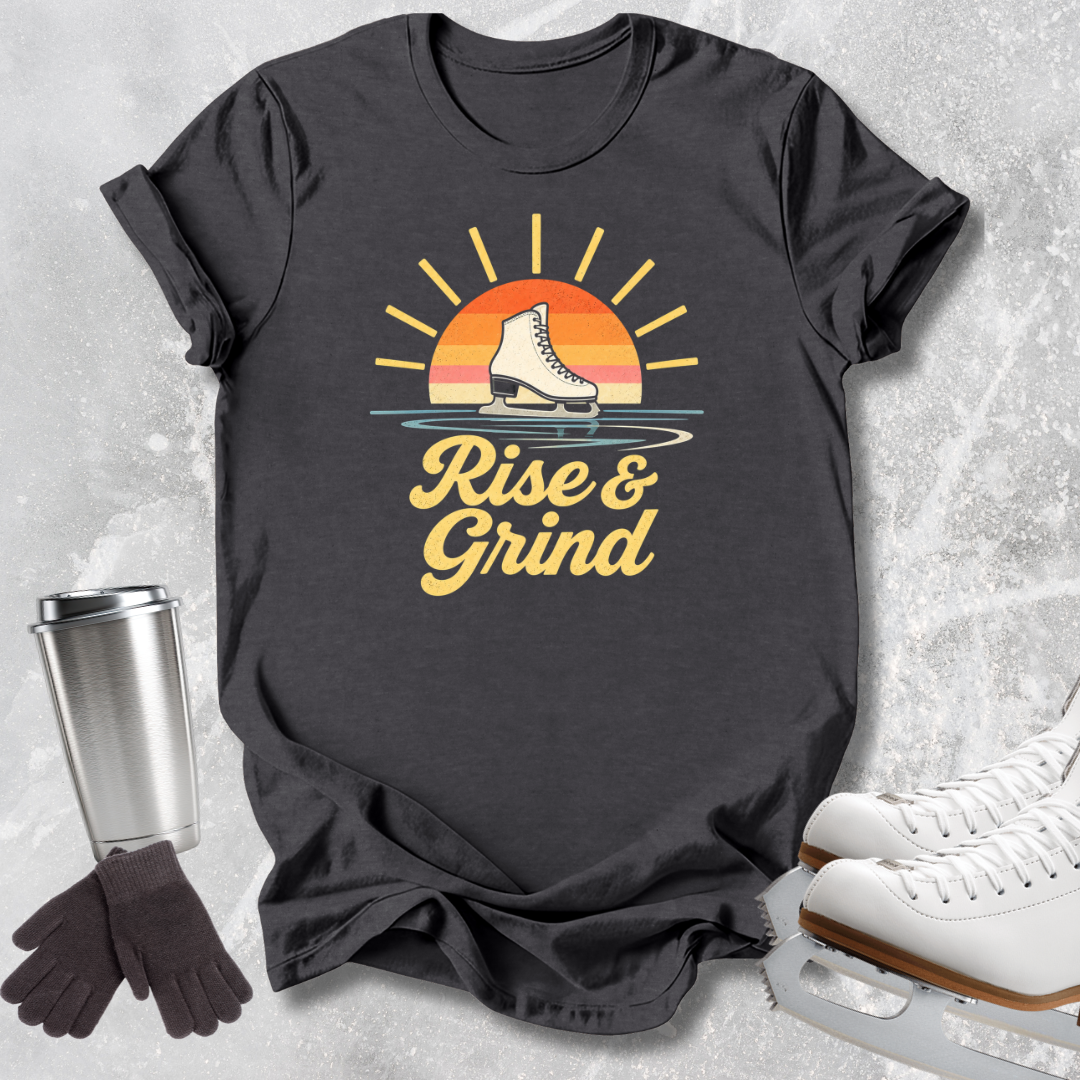 A dark charcoal gray graphic t-shirt featuring a retro-style sun graphic with a figure skating boot and the text 'Rise & Grind' in yellow. The shirt is styled in a flat-lay arrangement on a light gray textured background, accompanied by white ice skates, a stainless steel travel mug, and a pair of black gloves.