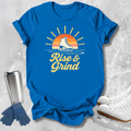 A bright blue graphic T-shirt laid flat on a light textured surface, featuring a retro sunset and ice skate design with the words 'Rise & Grind.' White ice skates, blue gloves, and a stainless steel travel mug are arranged around the shirt as styled props.