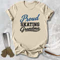 A beige graphic T-shirt laid flat on a light gray textured surface, with the words 'Proud Skating Grandma' printed on the front in blue and black. Ice skates, gloves, and a metal travel tumbler are arranged around the shirt as styling props.
