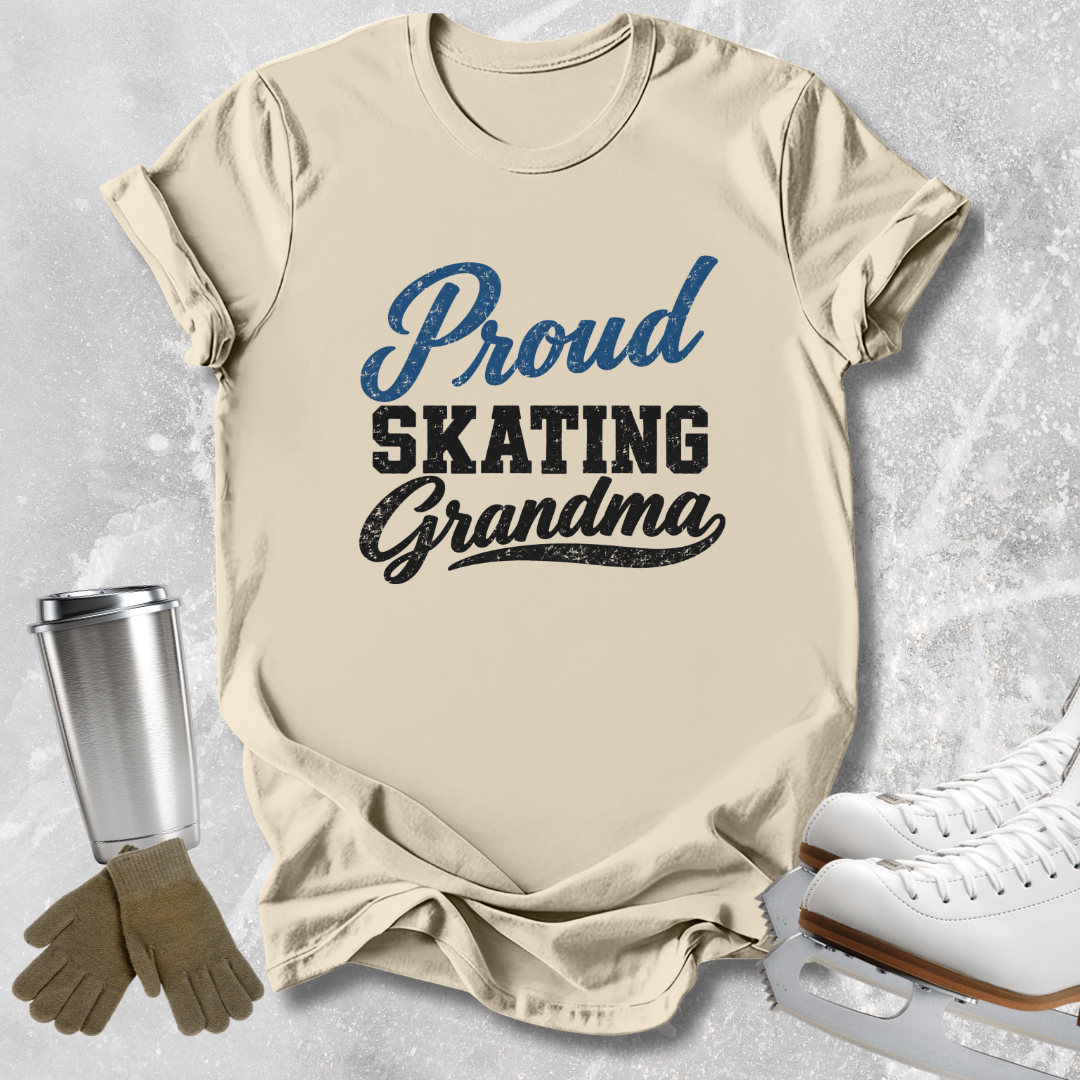 A beige graphic T-shirt laid flat on a light gray textured surface, with the words 'Proud Skating Grandma' printed on the front in blue and black. Ice skates, gloves, and a metal travel tumbler are arranged around the shirt as styling props.