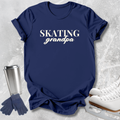 A navy blue graphic t-shirt with the words 'SKATING grandpa' printed in white, styled in a flat-lay with ice skates, gloves, and a travel tumbler on a light gray textured background.