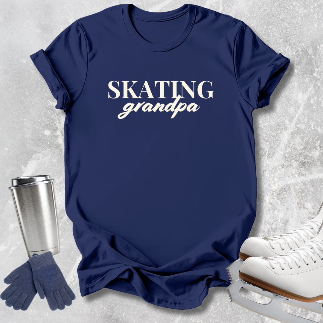 A navy blue graphic t-shirt with the words 'SKATING grandpa' printed in white, styled in a flat-lay with ice skates, gloves, and a travel tumbler on a light gray textured background.