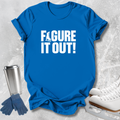 A bright blue short-sleeve T-shirt laid flat on a light gray textured surface, printed with the phrase 'FIGURE IT OUT!' in bold white letters. Ice skating props, including white ice skates, blue gloves, and a stainless steel travel mug, are arranged around the shirt.