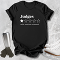 A black short-sleeve graphic T-shirt laid flat on a light textured surface, with white text reading 'Judges' above a one-star rating graphic and the phrase 'awful, would not recommend.' White ice skates, dark gloves, and a metal travel mug are placed around the shirt as styling props.