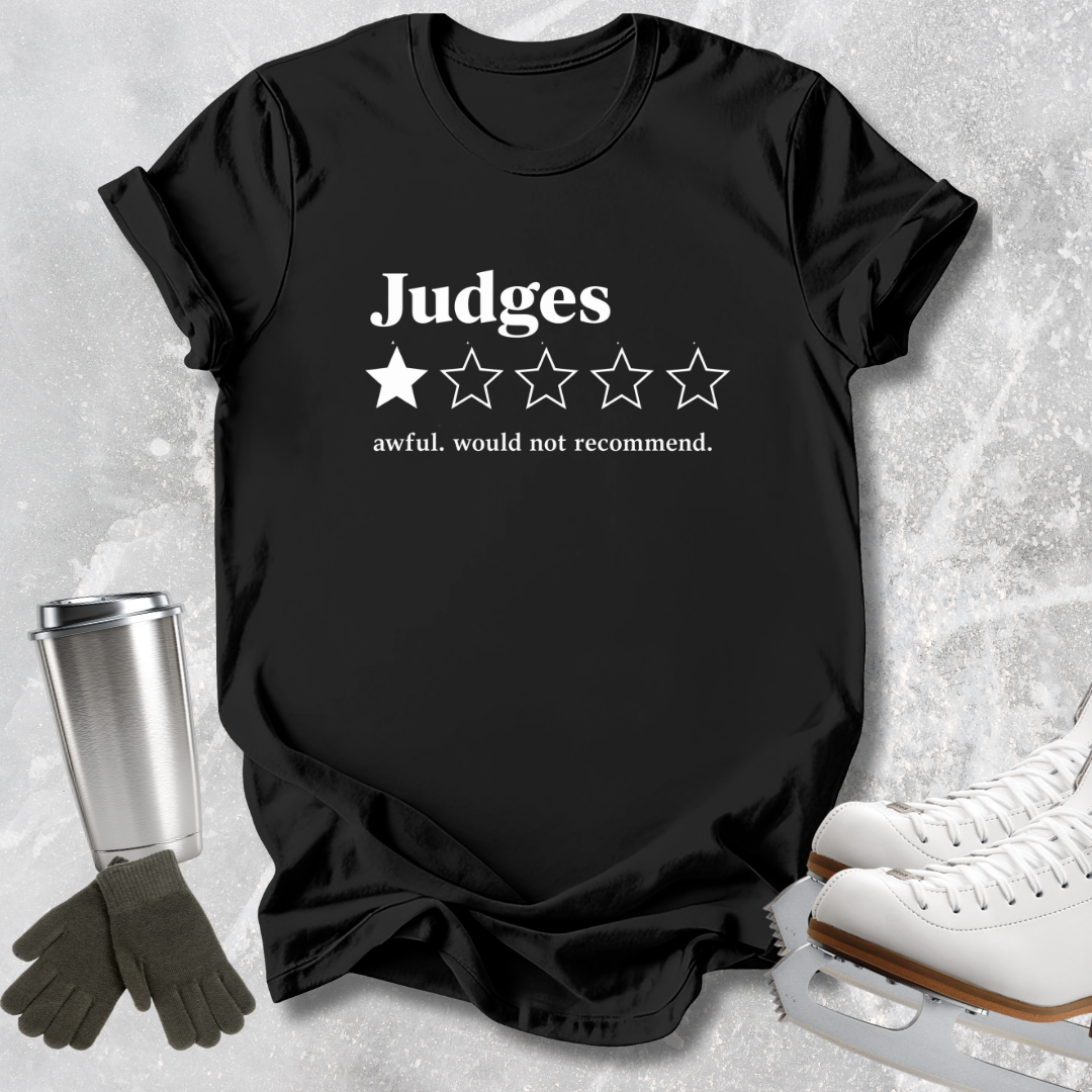 A black short-sleeve graphic T-shirt laid flat on a light textured surface, with white text reading 'Judges' above a one-star rating graphic and the phrase 'awful, would not recommend.' White ice skates, dark gloves, and a metal travel mug are placed around the shirt as styling props.