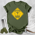 An olive green short-sleeve t-shirt with a yellow warning sign graphic that reads 'WARNING: EDGES AHEAD' with a figure skater silhouette. The shirt is styled in a flat-lay arrangement on a light gray textured background, accompanied by white ice skates, a silver travel mug, and green gloves.