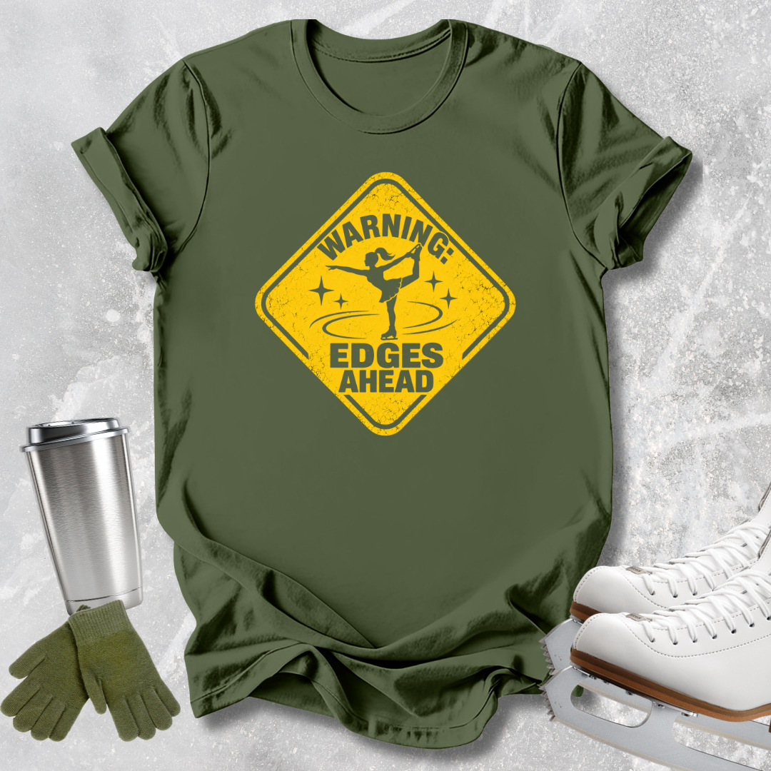 An olive green short-sleeve t-shirt with a yellow warning sign graphic that reads 'WARNING: EDGES AHEAD' with a figure skater silhouette. The shirt is styled in a flat-lay arrangement on a light gray textured background, accompanied by white ice skates, a silver travel mug, and green gloves.