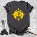A dark charcoal gray graphic t-shirt laid flat on a light gray textured surface, styled with ice skates, a metal travel tumbler, and a pair of gloves. The t-shirt features a yellow diamond-shaped road sign graphic with a figure skater and the text 'WARNING: EDGES AHEAD'.