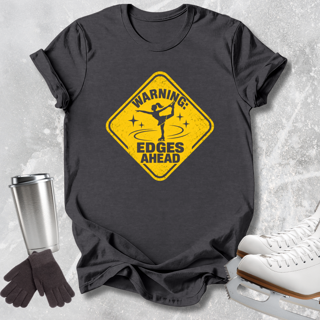 A dark charcoal gray graphic t-shirt laid flat on a light gray textured surface, styled with ice skates, a metal travel tumbler, and a pair of gloves. The t-shirt features a yellow diamond-shaped road sign graphic with a figure skater and the text 'WARNING: EDGES AHEAD'.