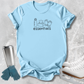 A light blue graphic t-shirt laid flat on a textured gray background, featuring a black line art design of winter essentials (water bottle, tissues, gloves, skates) with the word 'essentials' printed below. A stainless steel travel mug, blue gloves, and white ice skates are styled around the shirt.